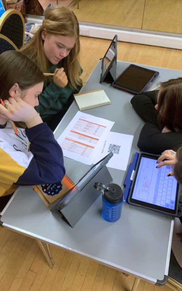 Students work together around paper and iPads on a desk.