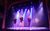 Students complete a practical task on stage. Washed in purple and blue light, three students pose in singing positions downstage. Upstage, a huddle of students watch them.