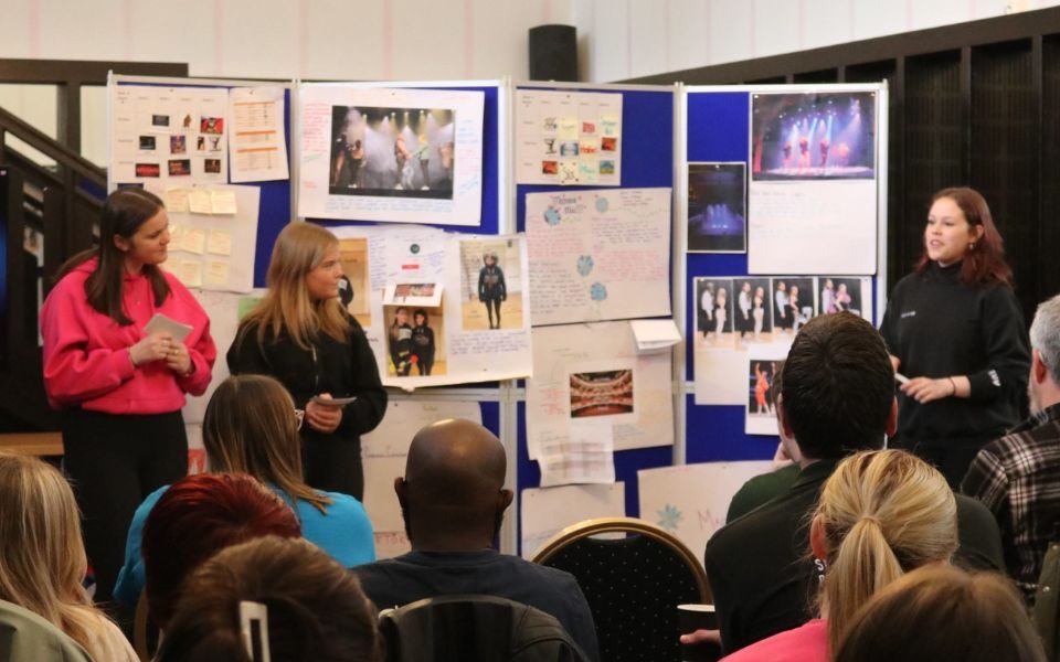 Accompanied by blue pinboards displaying their work, a group of students present to an audience of people.