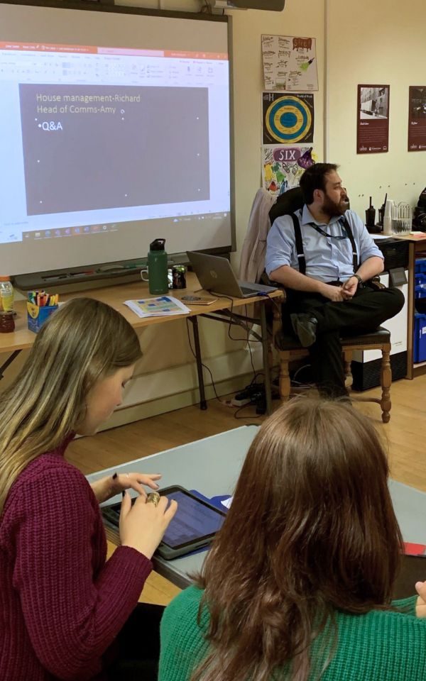 At the front of a classroom, Richard sits answering questions for sitting students, who are taking notes.