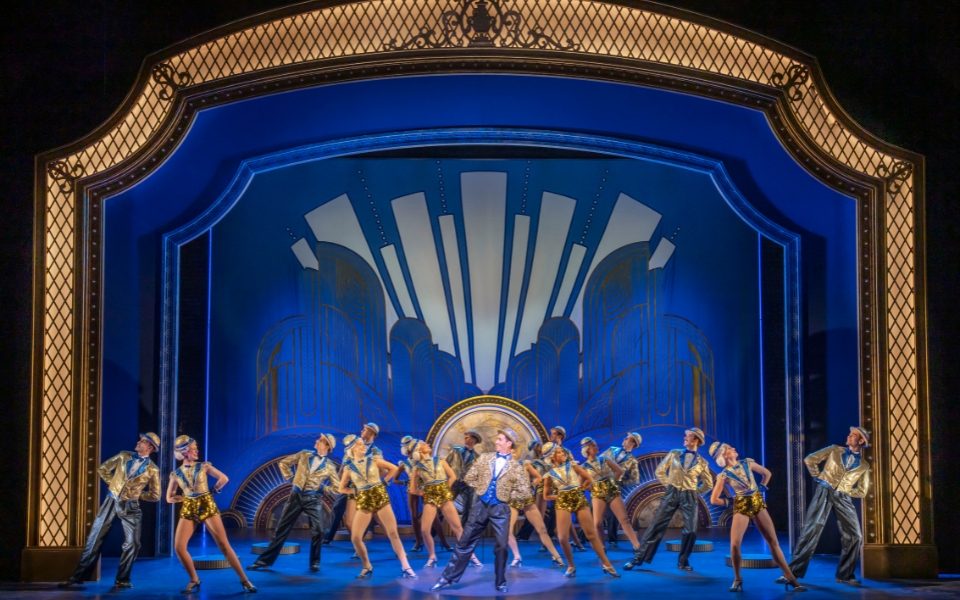 Dancers in formation facing each other in pairs and leaning away with their hands behind their backs. They stand in front of a blue backdrop.