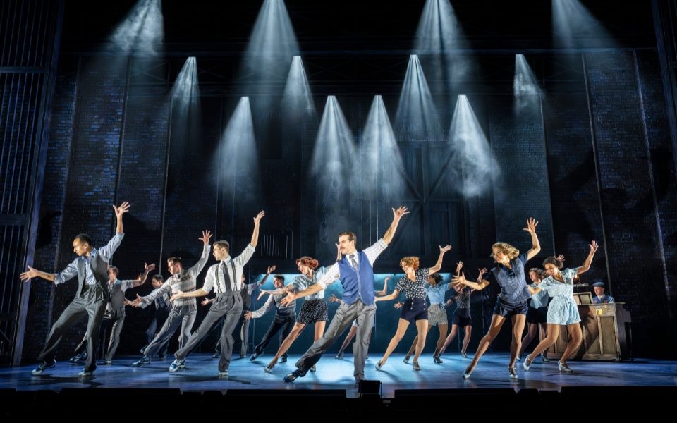 A chorus of dancers doing matching poses with the arms out while spotlights shine down on them.