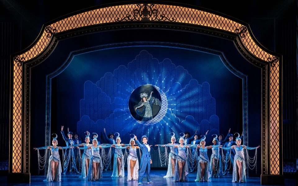 Dancers dressed in blue in front of a blue backdrop. There is a hole cut out of the backdrop with a woman standing on a moon.