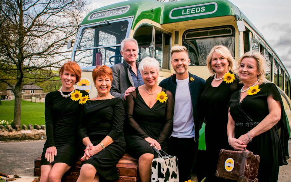 The 2018 cast of Calendar Girls the Musical with Gary Barlow and Tim Firth in front of a Leeds bus with luggage. They are wearing sunflowers on their green dresses.