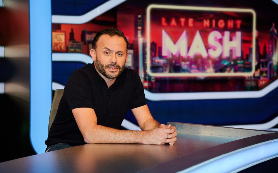 Geoff Norcott sits at a news desk with Late Night Mash on the screen behind him.