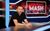 Geoff Norcott Geoff Norcott sits at a news desk with Late Night Mash on the screen behind him.