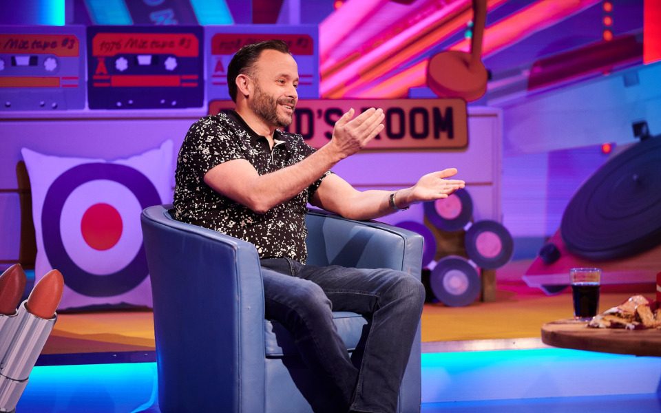 Geoff Norcott sits on a purple chair with a large record player and pieces of vinyls in the background.