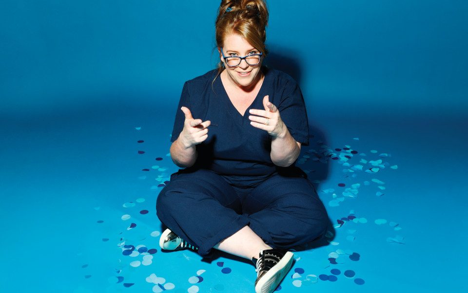 Georgie Carroll has her hair up and is wearing blue scrubs and points at the camera. The background is blue and full of blue confetti.