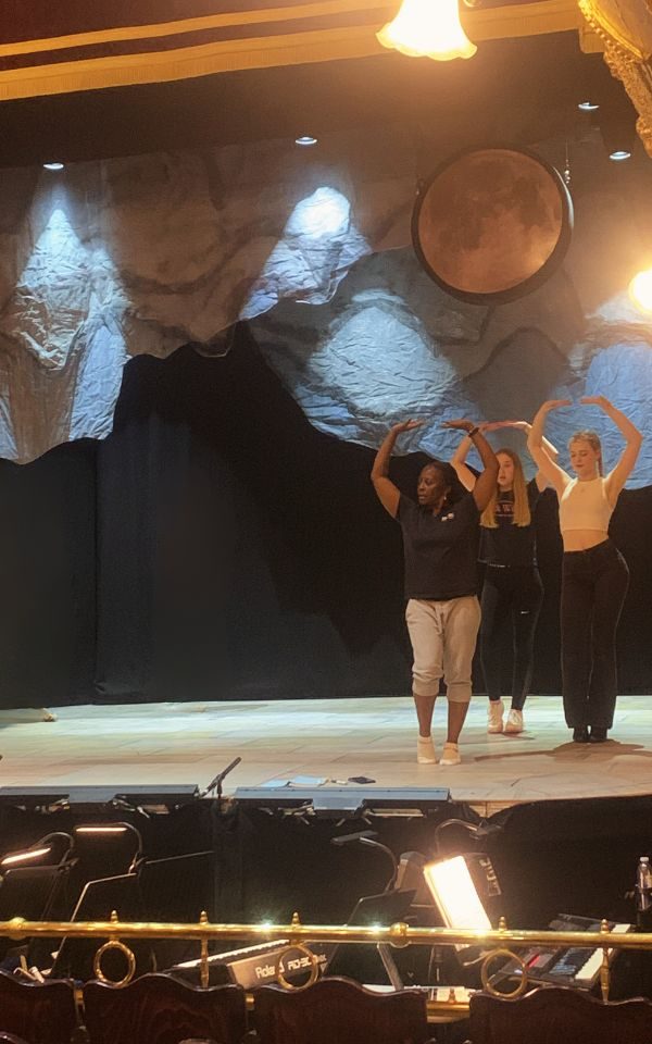 Ellie stands on stage as Morticia Addams, with another young person and Dawn (choreographer), standing with their arms in the air, mid-dance Credit: Kate Southam