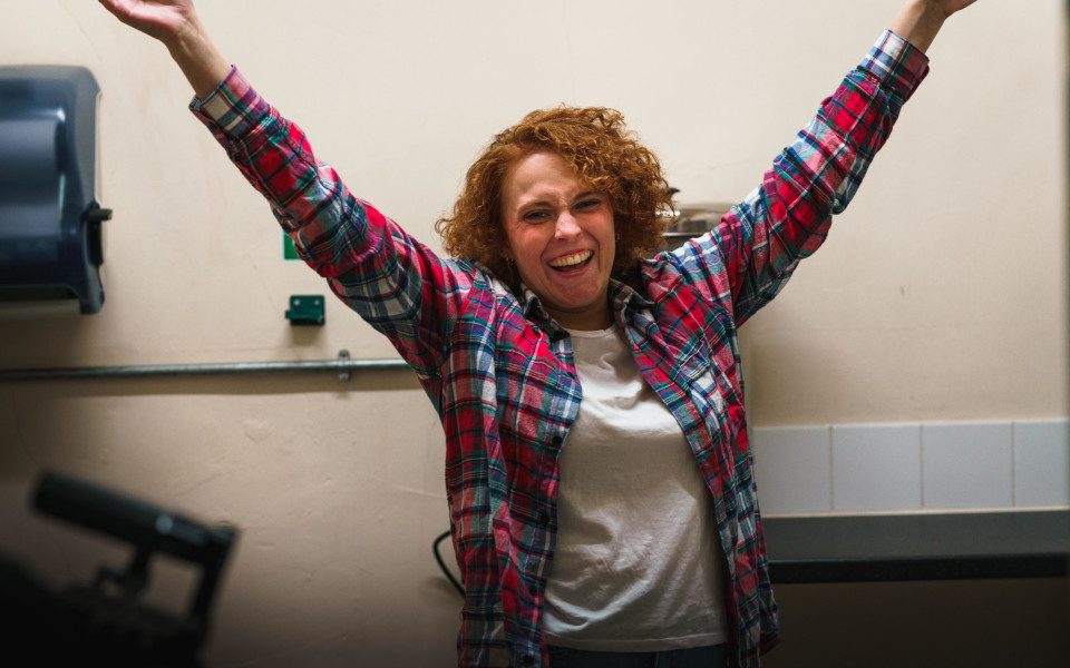 Tash is excitedly opening her arms and smiling. She is wearing a red plaid shirt.