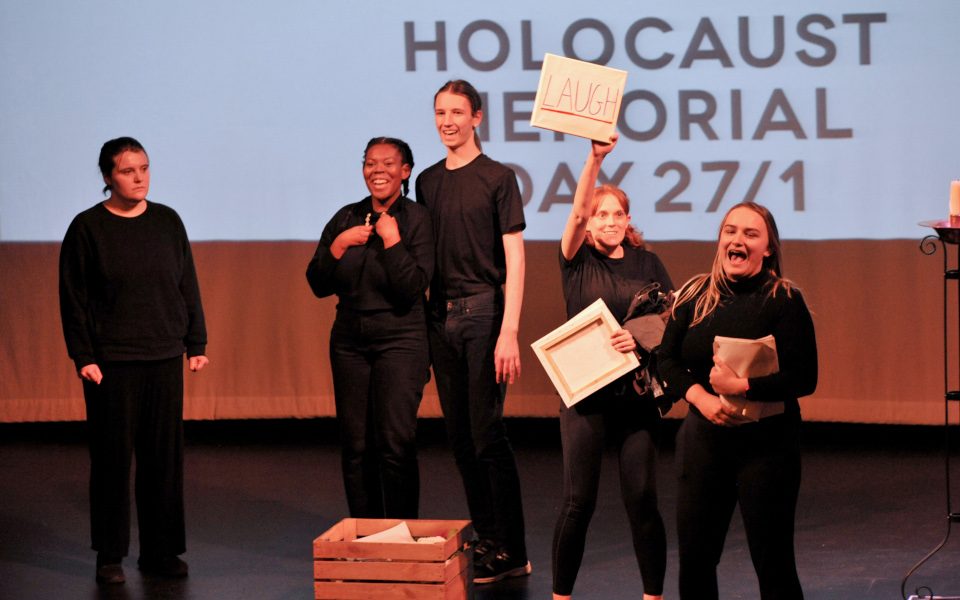 Five members of LAIT, including Tash Hudson, are dress in black. The screen behind them reads, Holocaust Memorial Day 27/1. Tash is holding a paper that reads Laugh.
