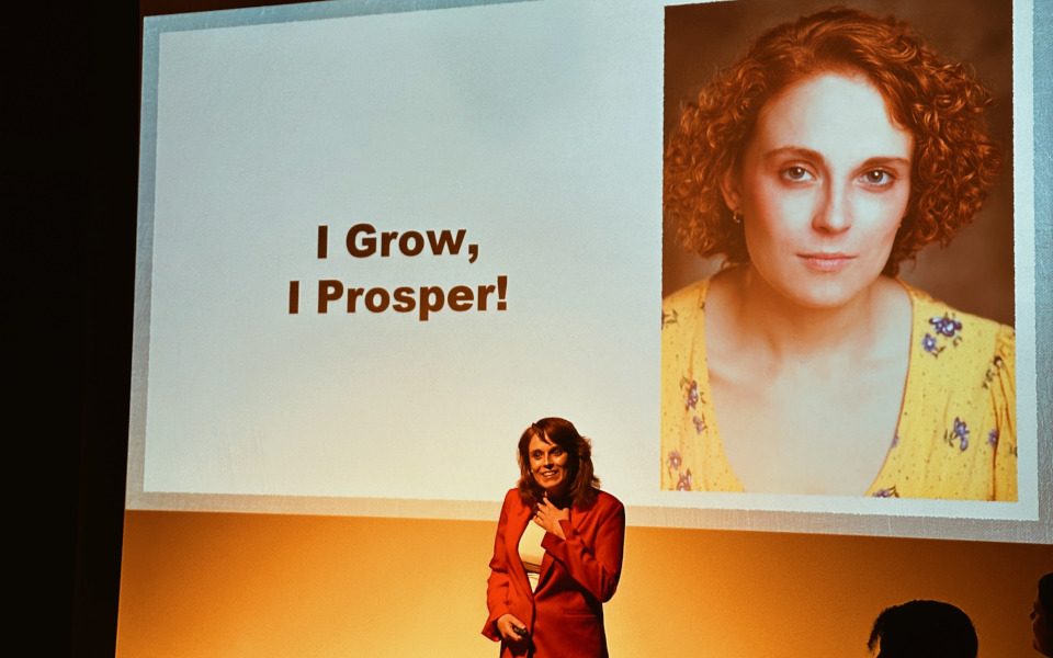 Tasha is standing in front of a screen of herself. The text reads, I grow, I prosper! She is wearing a red jacket.