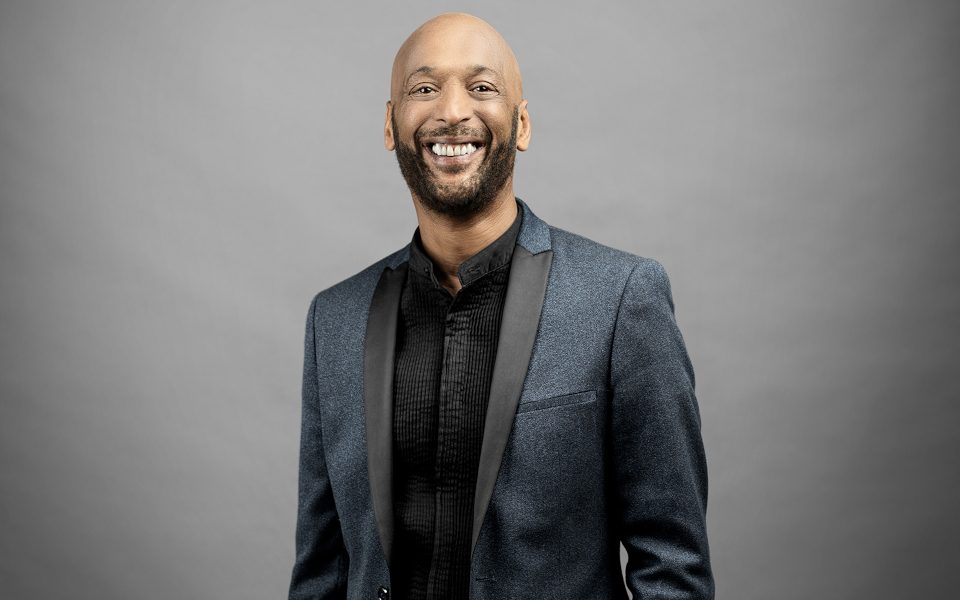 Tommy Blaize smiles while he wears a blue suit and a black shirt. He is standing with his left hand in his pocket in front of a grey background.
