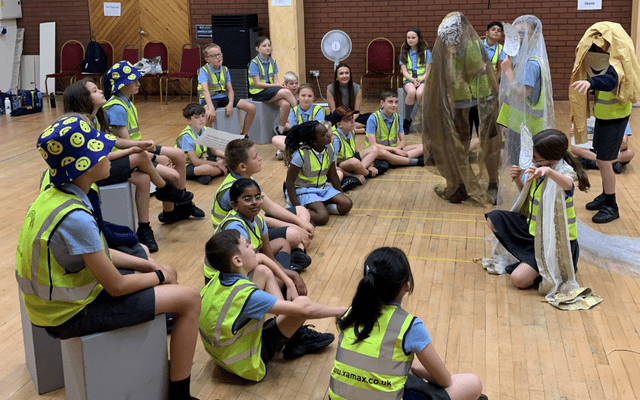 Primary school students have made an amphitheatre by sitting on stools, some participants are wearing masks and fabric pretending to be actors on stage.
