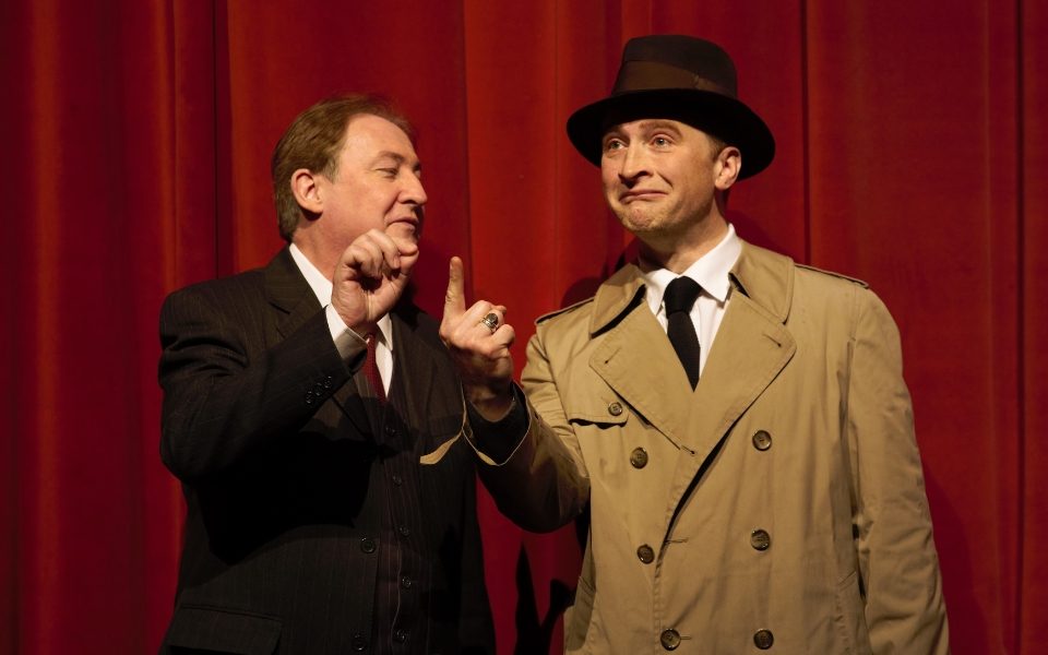 A man in a trenchcoat and hat points a finger up in the air next to a man in a suit. They are in front of a red curtain backdrop.
