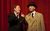 Hancock's Half Hour A man in a trenchcoat and hat points a finger up in the air next to a man in a suit. They are in front of a red curtain backdrop.