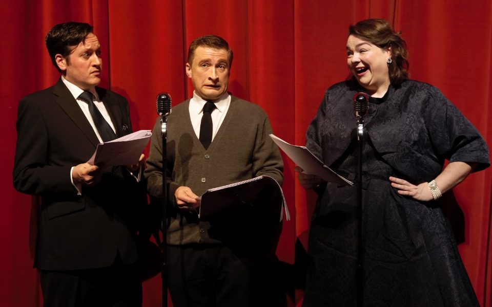 Two men and a woman hold scripts and pull faces around a microphone. They wear smart clothes and stand in front of a red curtain backdrop.