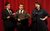 Hancock's Half Hour Two men and a woman hold scripts and pull faces around a microphone. They wear smart clothes and stand in front of a red curtain backdrop.