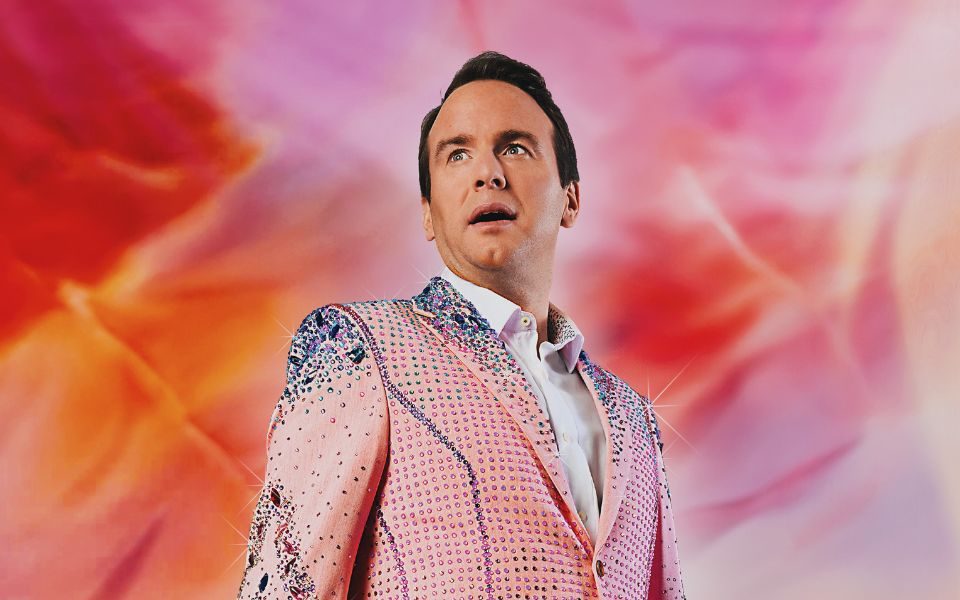 Matt Forde wearing a pale pink bejewelled suit with blue details and white shirt underneath. He is against a pink and orange wispy background. The image is mimicking Taylor Swift's Eras Tour artwork.