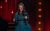 Rosie Jones Rosie Jones in a teal dress performing in a microphone on a stage draped in red curtains