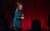 Rosie Jones Rosie Jones in a teal dress performing in a microphone on a stage draped in red curtains