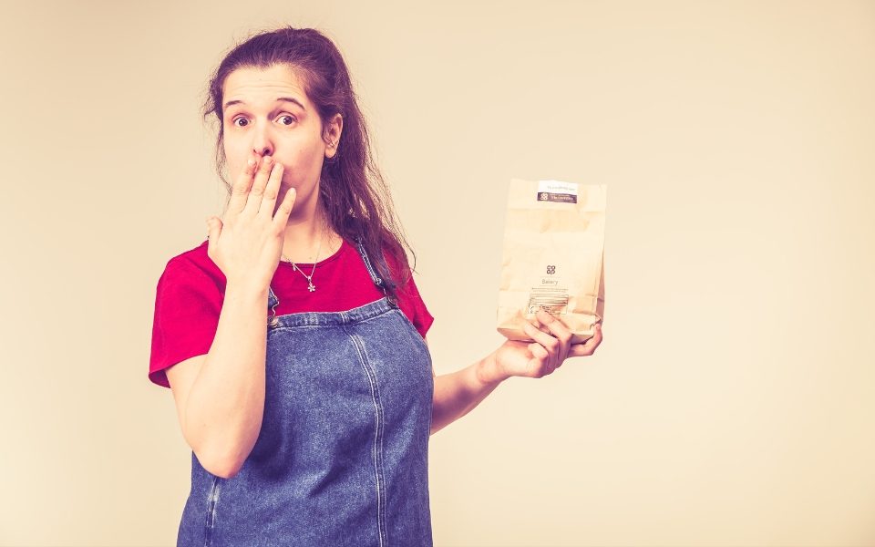 Rosie Jones covering her mouth with her hand and looking at the camera while she holds a bakery packet in the other hand