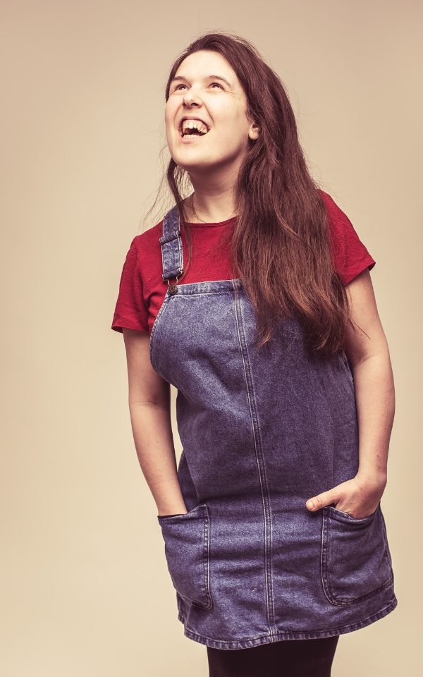 Rosie Jones in a denim dress looking up and laughing with her hands in her pockets