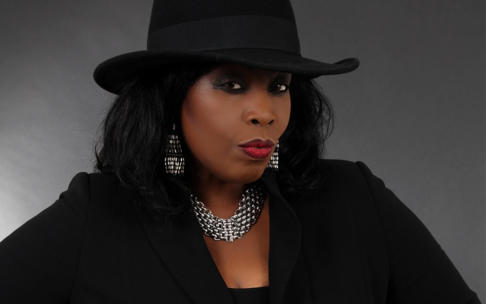 Ruby Turner is wearing a black cowboy hat along with silver earrings and a chunky necklace. Her smoky eye makeup is paired with a bold red lip. Ruby is wearing a black shirt and blazer.
