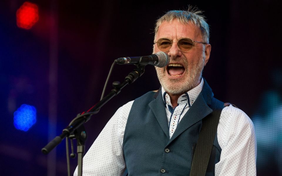 Steve Harley sings into a microphone on stage.