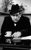 Tony Hancock A black and white photo of Tony Hancock sat at a desk in a suit and hat with a cup of tea next to his hand.