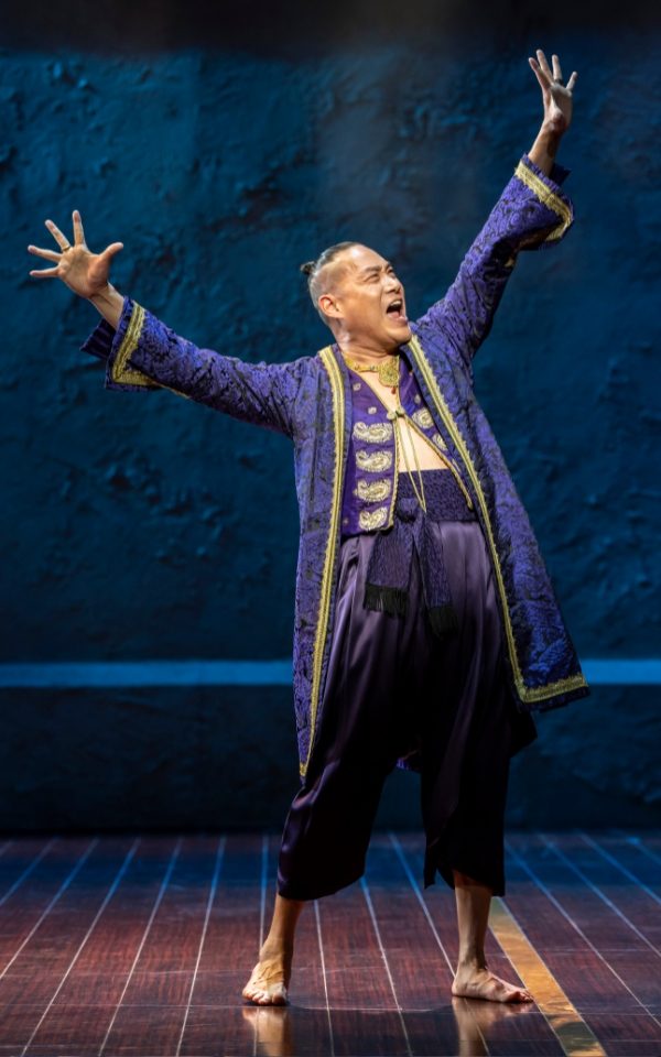 Darren Lee as the King of Siam in a dark blue traditional Thai jacket and trousers. He stands barefoot with his arms out above his head.