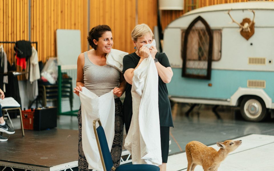 Helen Évora as Meg Page and Louise Winter as Mistress Quickly in rehearsal for Falstaff. They are holding sheets and standing next to a deer. There is a caravan in the background.