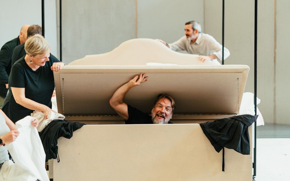 Henry Waddington rehearsing the role of Falstaff with Opera North. He is inside a chest with an actor helping him open it. There are people working in the background.