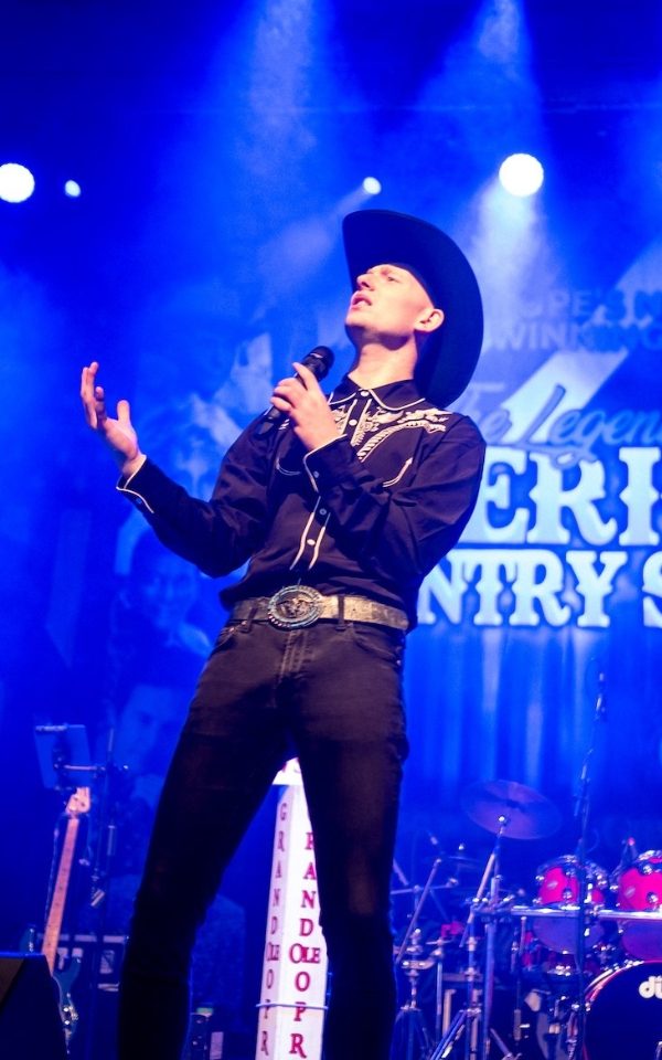 Kelan Browne in a country-style shirt and cowboy hat singing into a handheld microphone in front of the Legends of American Country logo backdrop