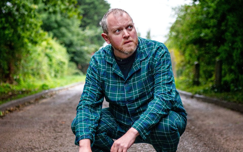 Miles Jupp is standing on a path surrounded by trees. He is wearing green pyjamas and no shoes. He is looking off to the side.
