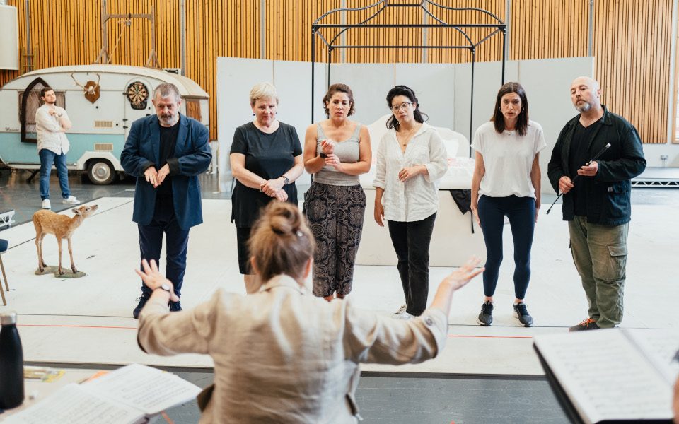 Olivia Fuchs directing the cast in rehearsal for Falstaff. There is a bed, a caravan, and a deer in the background.