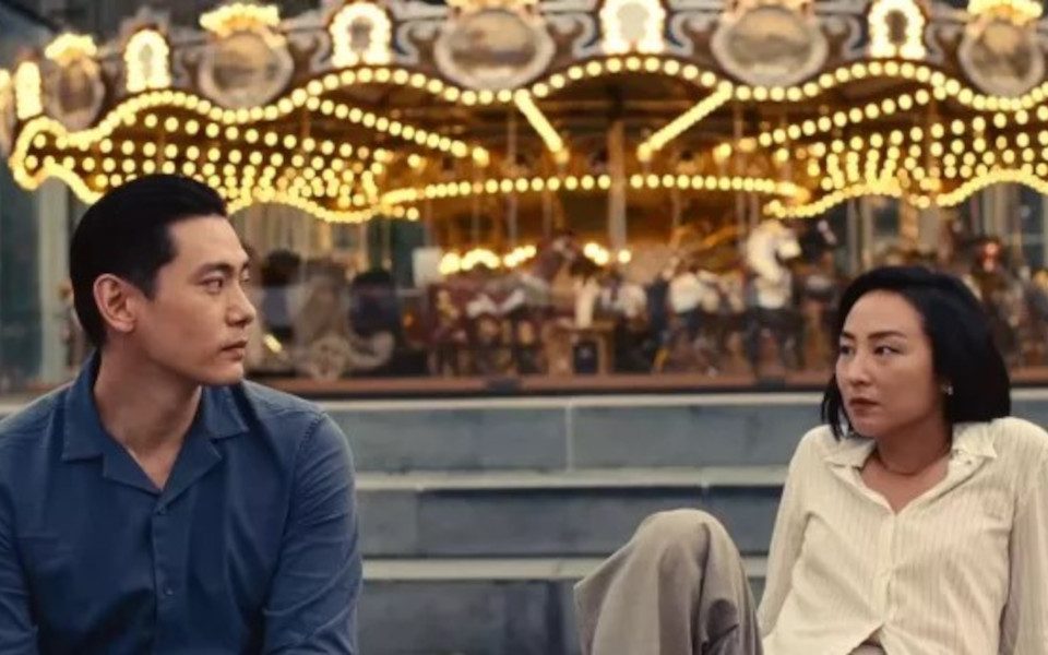 A still from Past Lives. Two characters are sitting in front of a carousel talking.