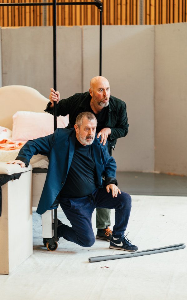 Richard Burkhard (back) as Ford with Paul Nilon (front) as Dr Caius. They are crouching in front of a bed and looking at something in the distance.