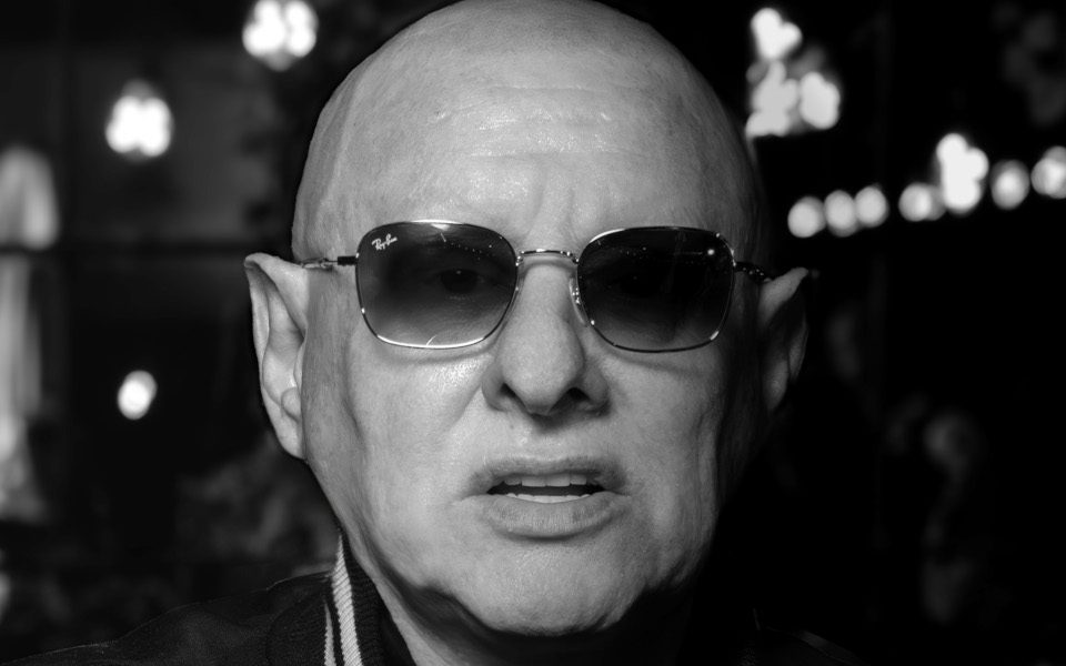 A close-up of Shaun Ryder. He is wearing sunglasses and a black jacket.