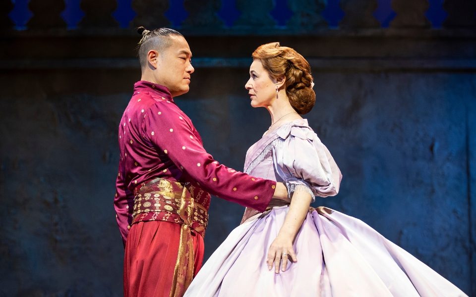 Darren Lee in a red traditional Thai costume holding Annalene Beechey at the waist as they look into each others' eyes. She wears a lilac ballgown.