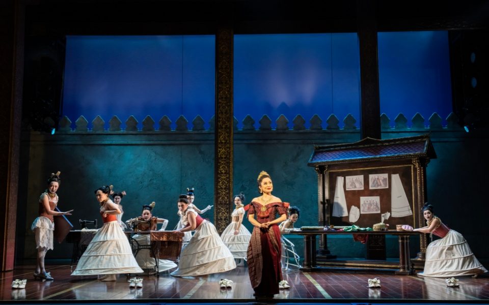 Cezarah Bonner standing centre stage in red traditional Thai dress and a crown. Behind her the ensemble wear long skirts in a classroom scene.