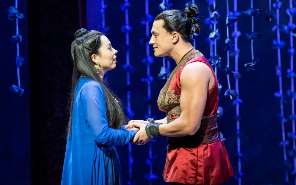 Marienella Phillips and Dean John-Wilson in The King and I. The hold hands and look into each others' eyes. She wears a blue robe and he wears traditional red Thai clothing.