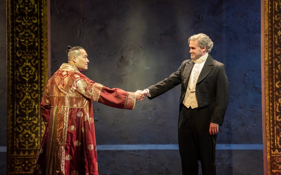 Darren Lee and Sam Jenkins-Shaw in The King and I. Darren wears a red and gold robe and shakes hands with Sam in a black suit and bowtie.