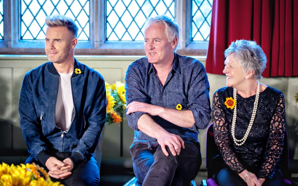 Gary Barlow, Tim Firth, and a Calendar Girl sit together with sunflowers.