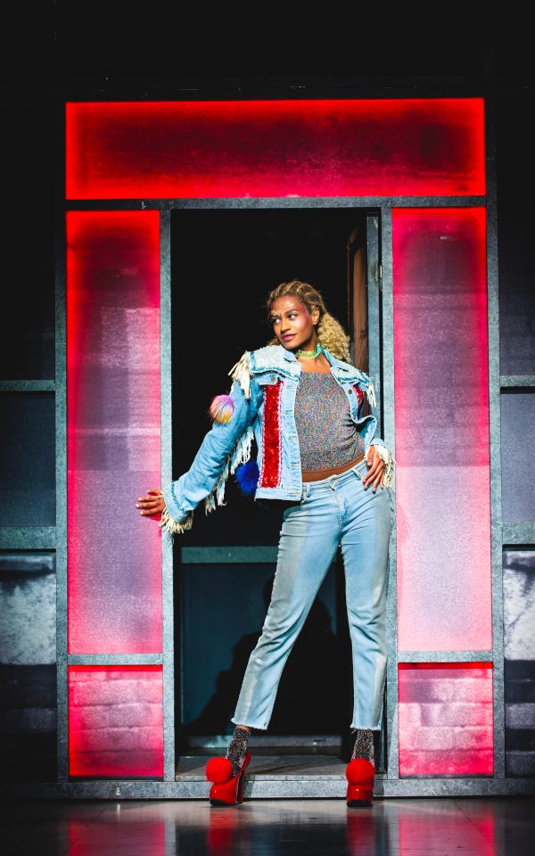 Jamie is wearing a jean jacket and jeans. He has his hair up and is wearing bright red heels. He is walking out of a red-lit doorframe.