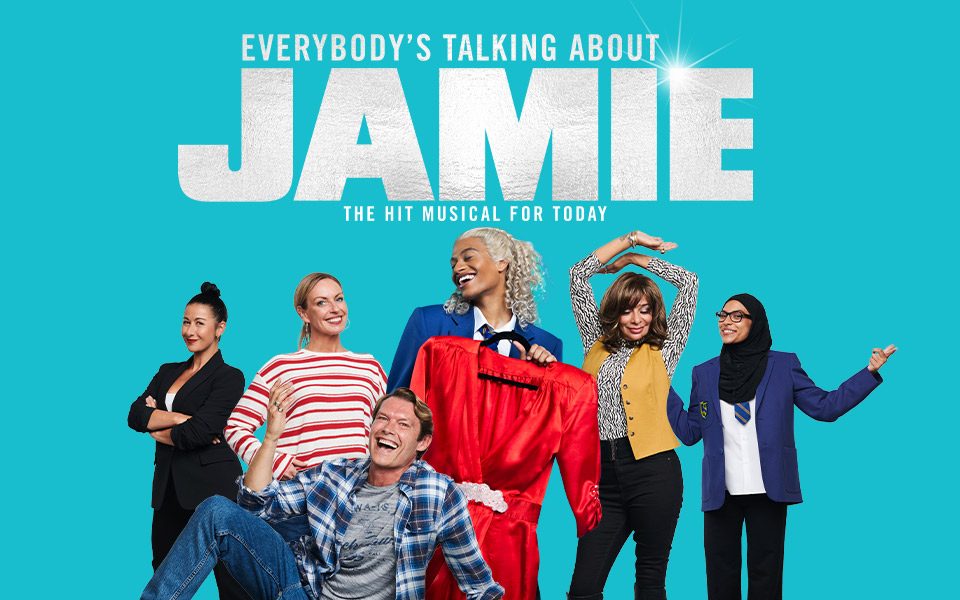Everybody's Talking About Jamie: The Hit Musical For Today in white writing with a shining spot on the E. Ivano Turco as Jamie. Ivano is holding a red dress and standing with the rest of the cast.