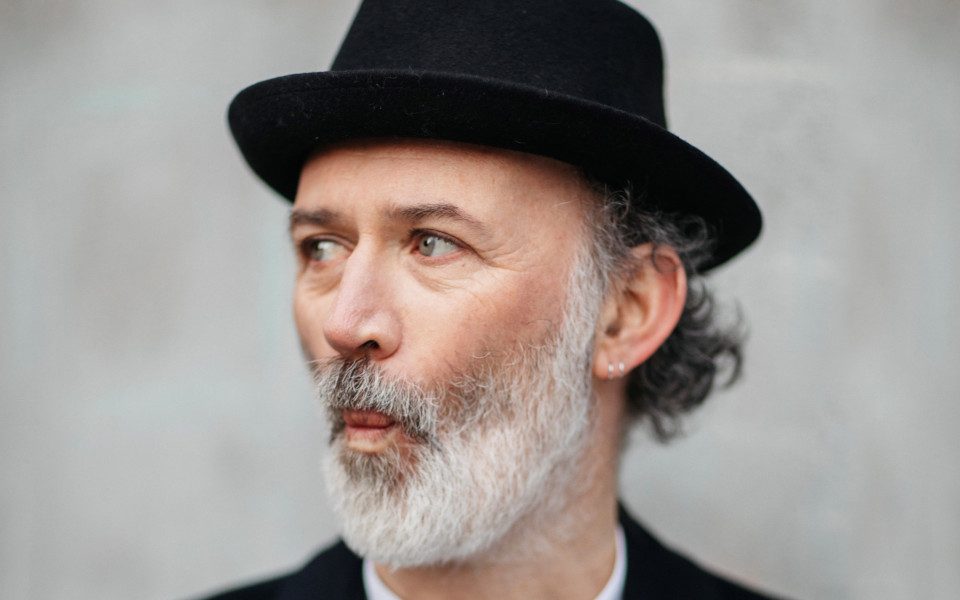 Tommy Tiernan is wearing a black fedora and jacket. He has a white beard and is pursing his lips.