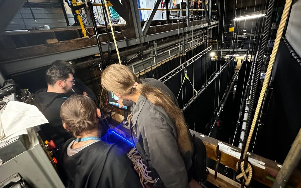 Three people gathered around a screen on the fly platform backstage of City Varieties. They look down on the black tabs that are flown on and off stage.