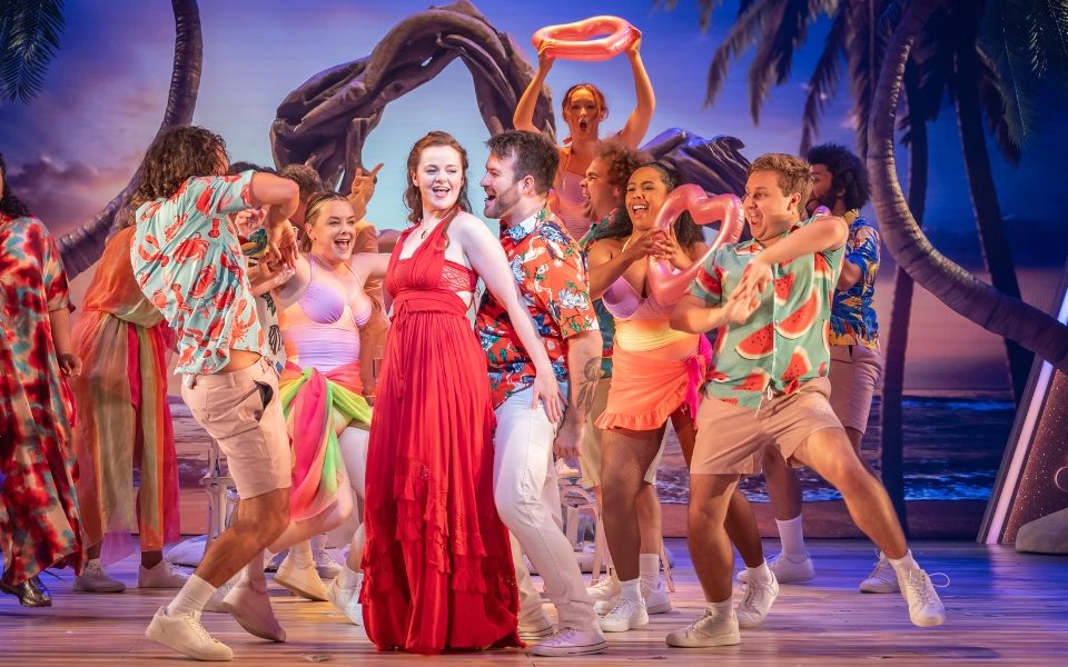 A woman in a red dress with a man dancing behind her in a floral shirt. The rest of the cast dance behind in Hawaiian shirts.