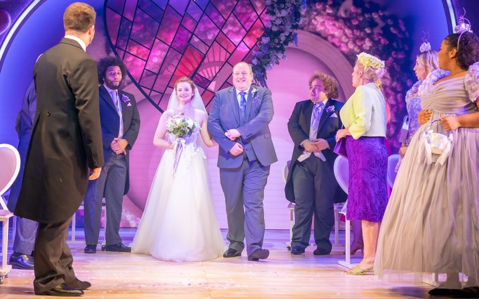 A bride in a wedding dress walking down the aisle with a man in a suit surrounded by wedding guests.